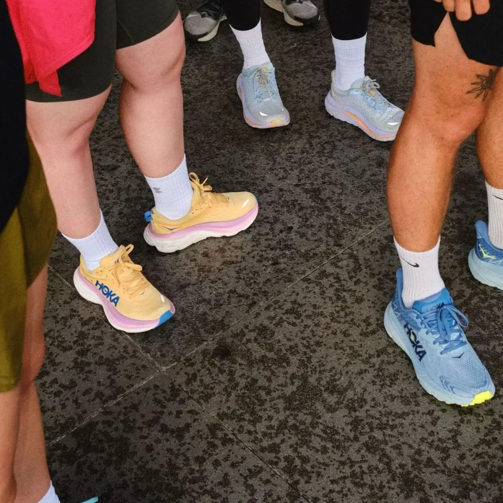 A group of people from a running club stands on a textured surface, all wearing athletic shoes—mainly Hoka—in various colors. Only their legs and feet are visible, showing their love running together.